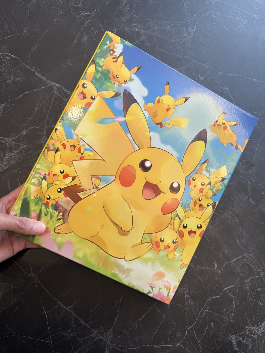 A hand holding a card binder whose cover shows many Pikachu