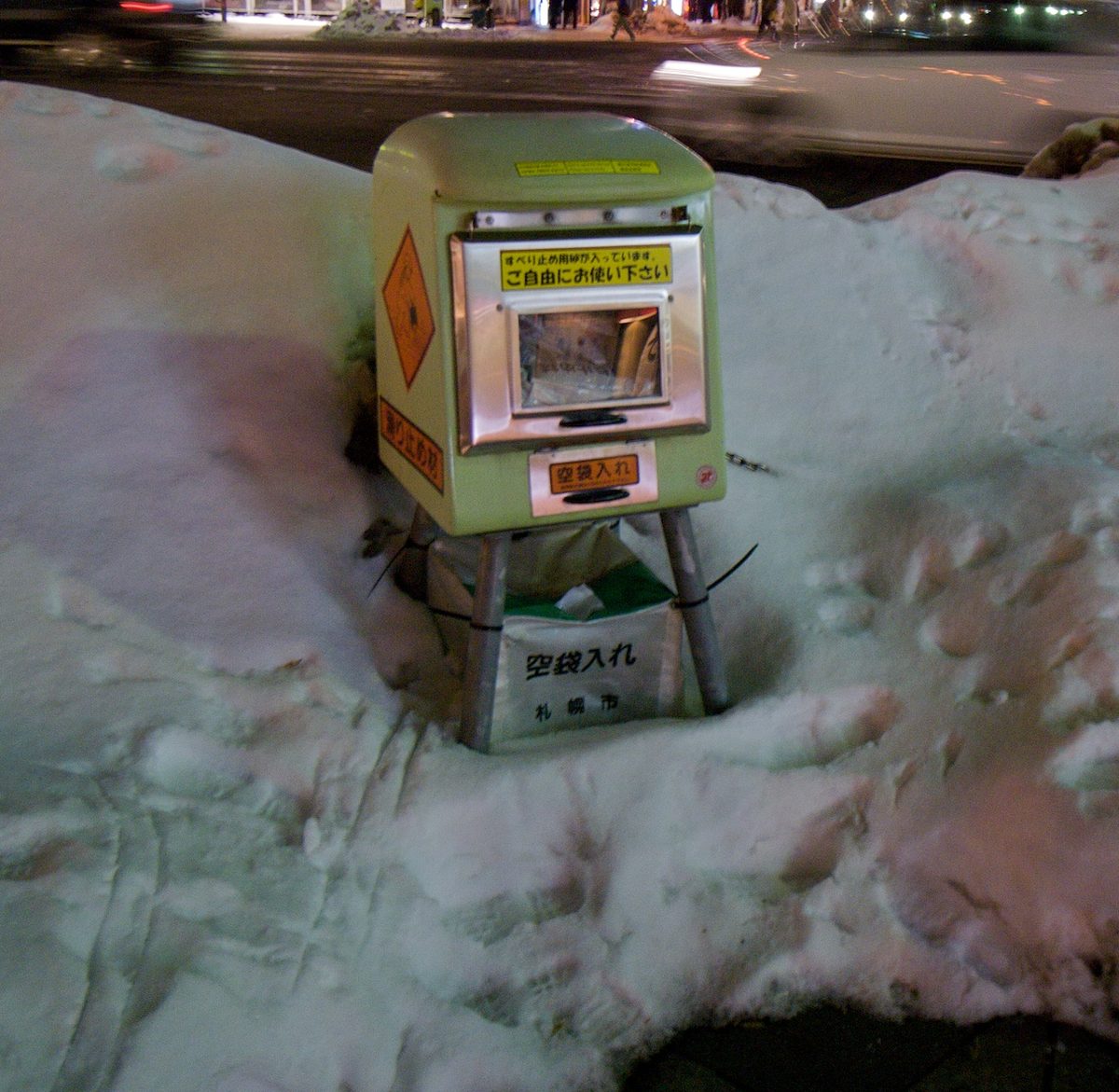 A small box of anti-slip sand standing in the snow beside a city street at night