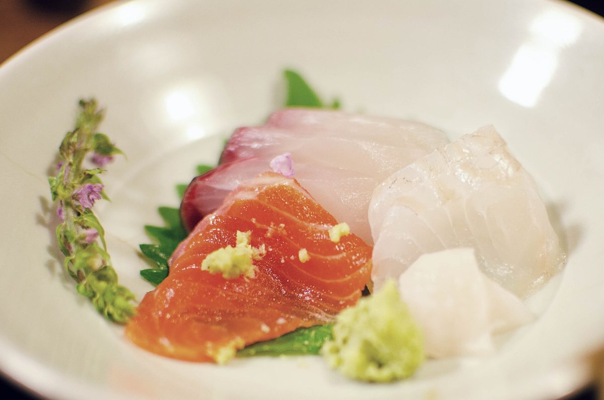 A plate of sashimi: slices of salmon and white fish on green leaves, a small mound of grated wasabi, a little wasabi on the salmon, and a sprig of shiso flowers