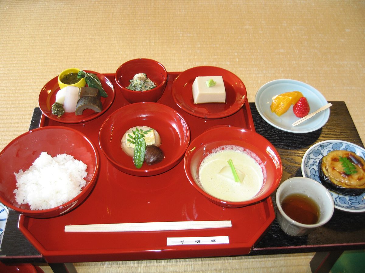 A shojin ryori meal on a red lacquered tray: rice, sesame tofu, simmered vegetables and small side dishes, with chopsticks laid across the front