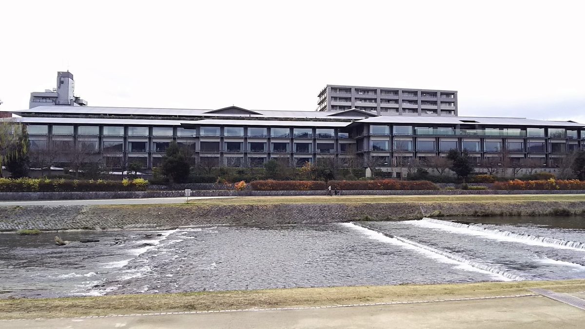 A long, low hotel building with dark wooden lines along the bank of the Kamo River, seen across the shallow water