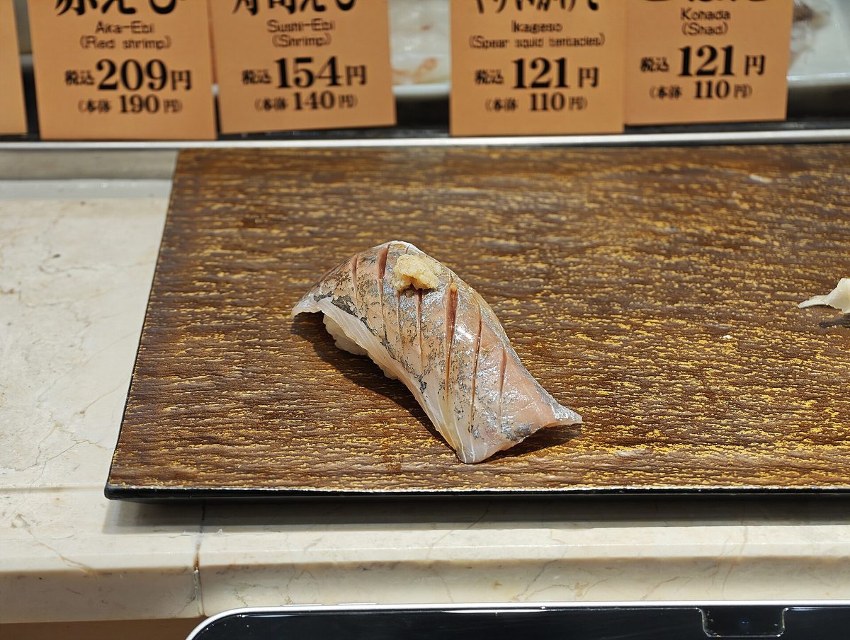 A piece of horse mackerel (aji) nigiri with silver skin on a dark tray at a sushi counter