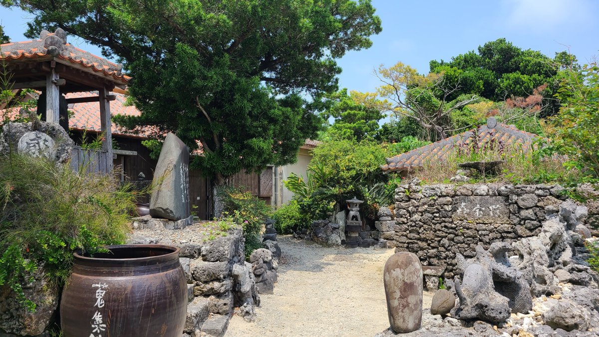 A sandy path between walls of grey coral stone, with a large clay jar, stone posts and a red-tiled roof behind the trees