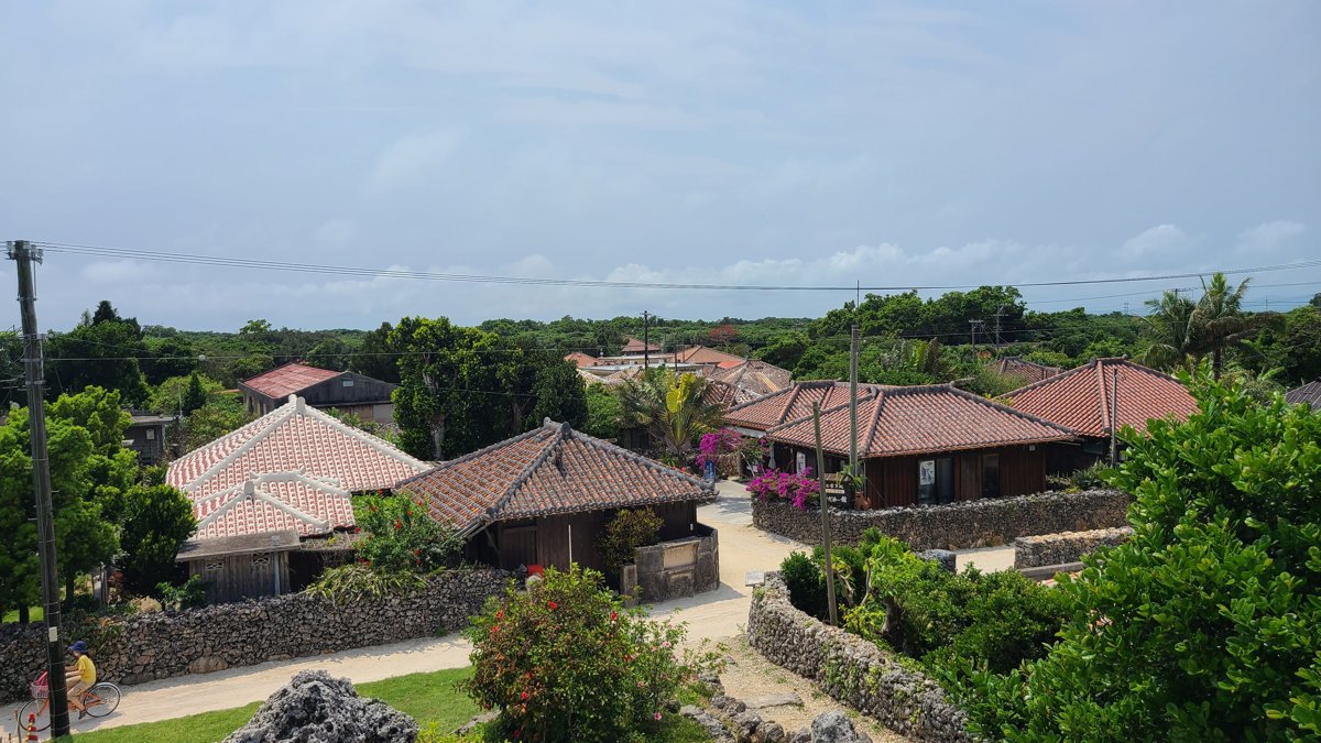 Red-tiled roofs of low houses among green trees and coral stone walls, with pink bougainvillea, under a cloudy sky on Taketomi Island