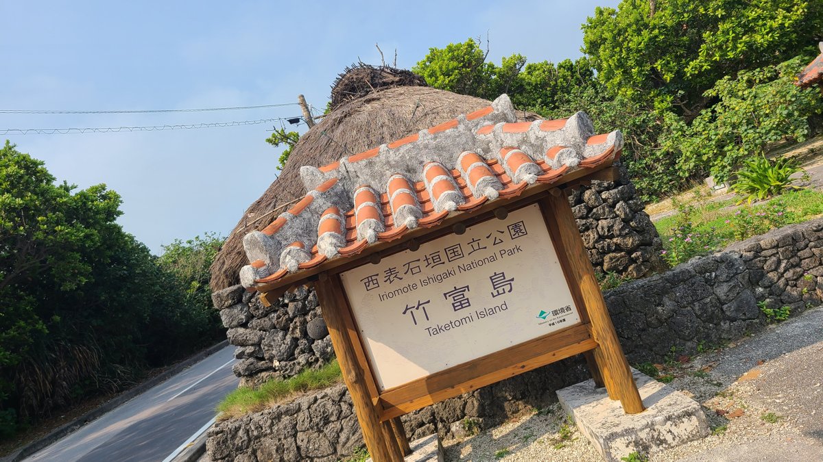 A wooden sign reading Taketomi Island under a small red-tiled roof, beside a stone wall and a road lined with trees