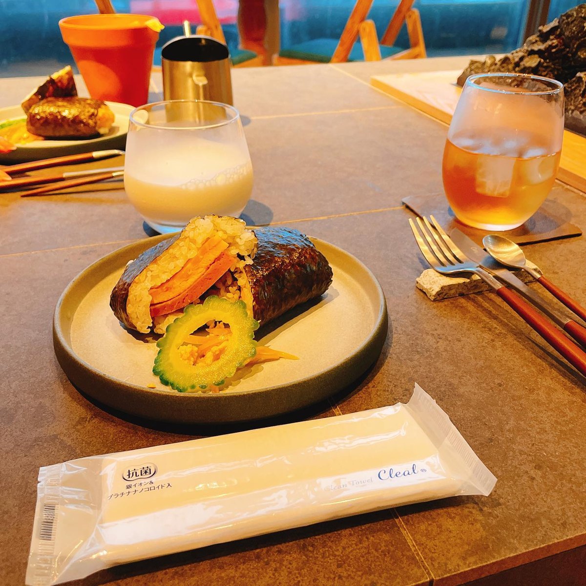 A rice ball wrapped in seaweed with slices of luncheon meat and egg inside and a ring of bitter melon on a plate, with a glass of smoothie and a glass of iced tea on the table