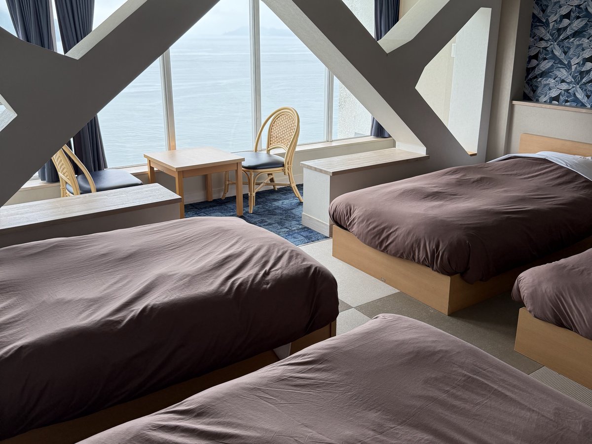 A bright guest room with several beds, white beams and large windows looking out to sea