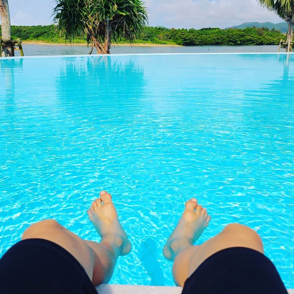 Bare feet dangling into a bright turquoise pool that looks out to green hills and palm trees