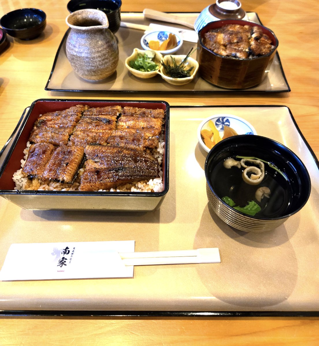 In front, unaju: grilled eel on rice in a square lacquered box, with a clear soup and pickles. Behind it, hitsumabushi in a wooden tub with green onion, nori and wasabi and a pot of broth