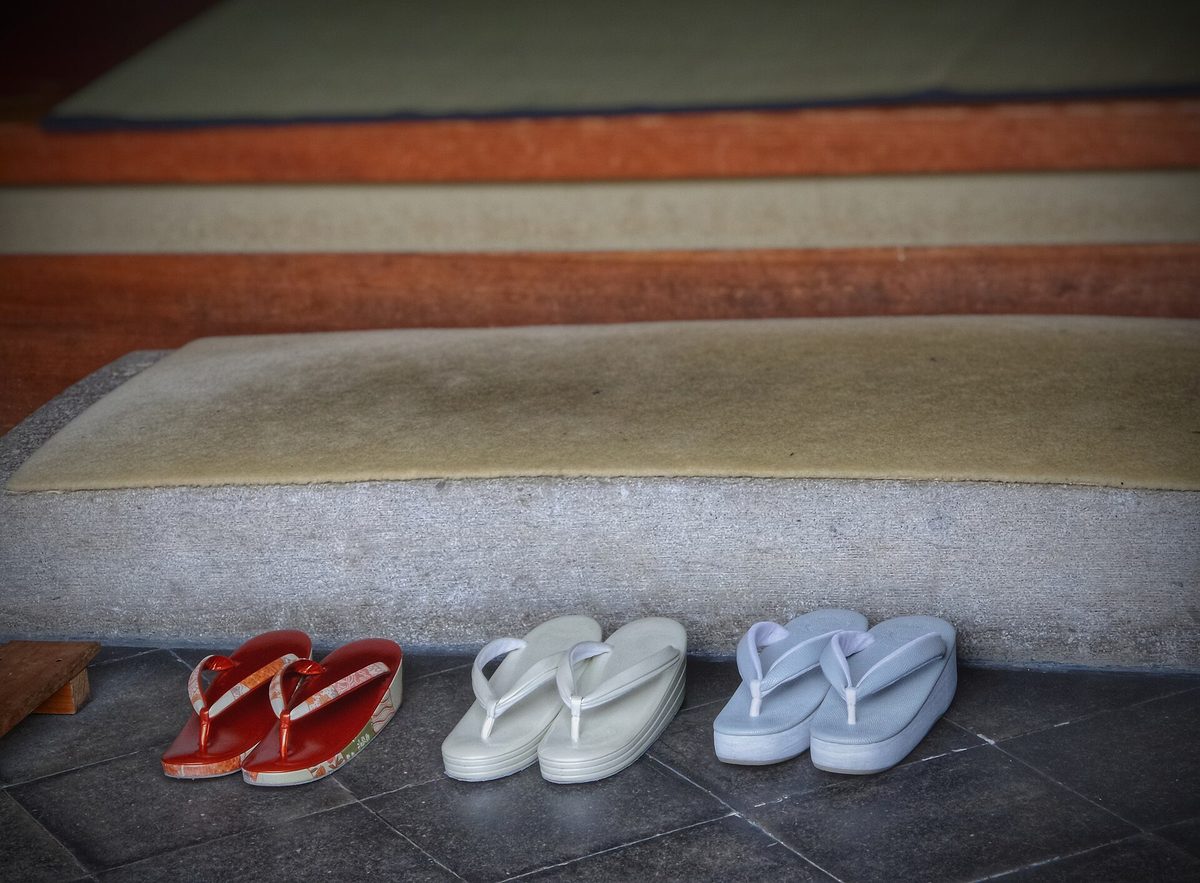 Three pairs of zori sandals lined up on a stone floor below the raised step of a traditional Japanese entrance