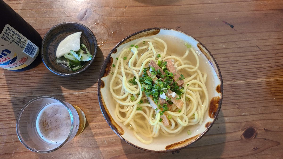 A bowl of soba noodles topped with chopped green onion and small pieces of pork, on a wooden table with a glass of water
